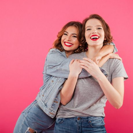 Image of happy two women friends standing isolated over pink background. Looking camera hugging.